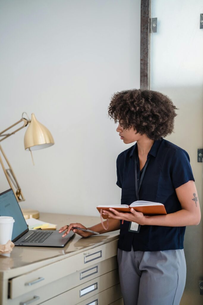 A woman holding a notebook and using a laptop.